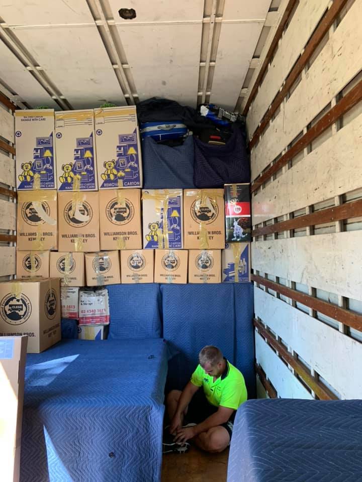 Removalist kneeling to secure blanket-wrapped furniture below stacked boxes in a truck
